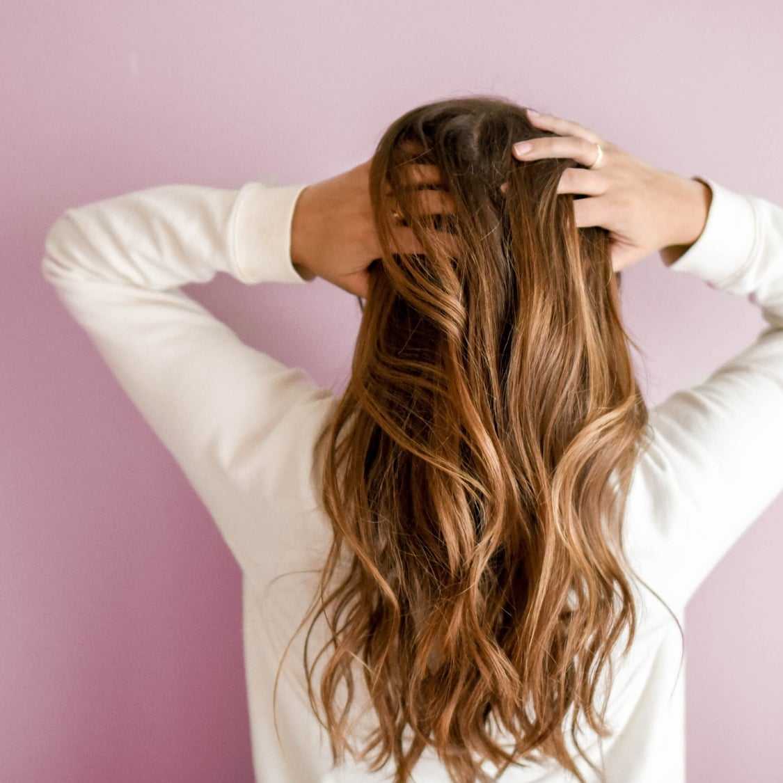 Woman with long hair facing away, hands in hair, pink background.